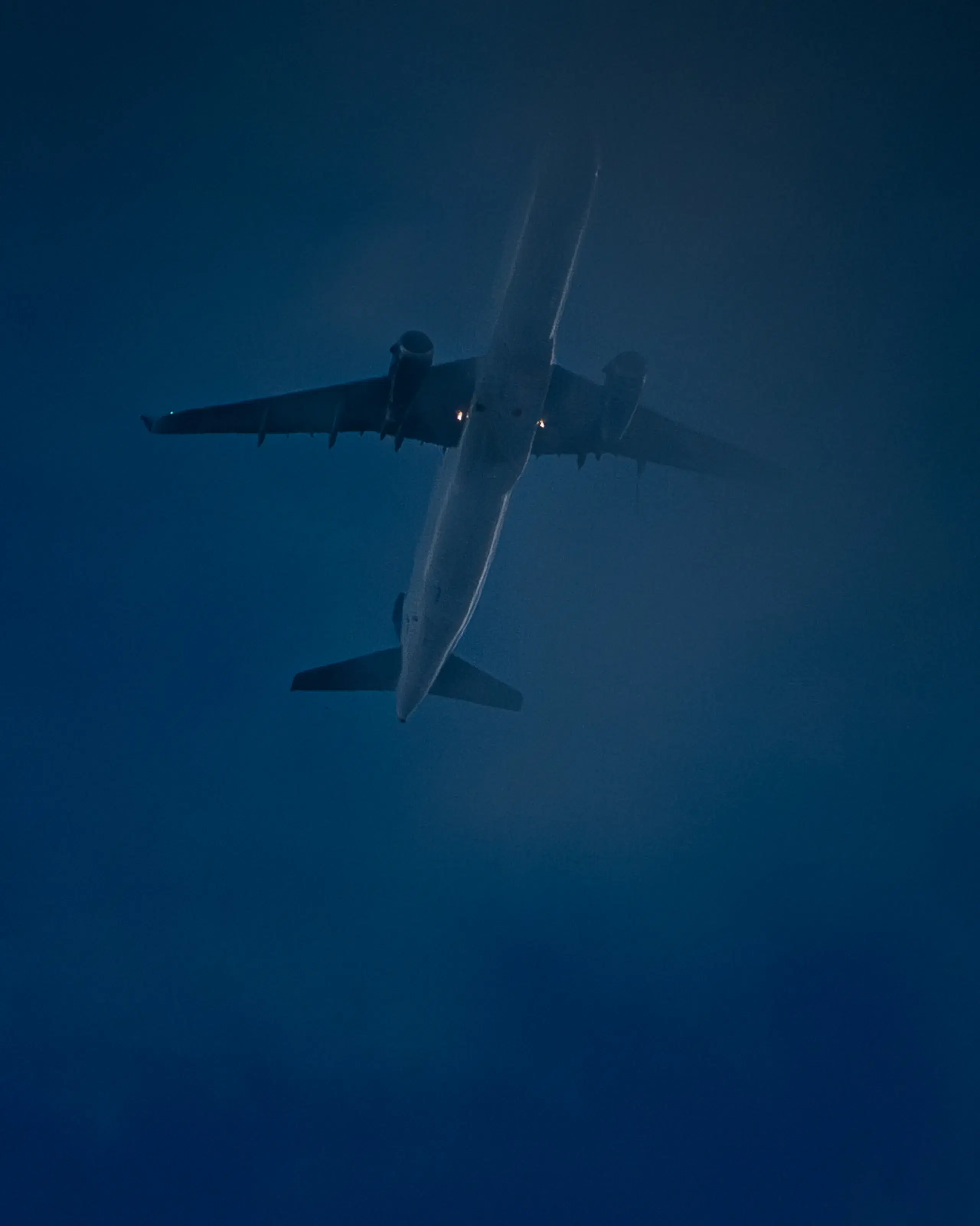 An airliner on approach, seen from below through dense blue haze that flattens it almost to a silhouette.